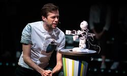 A white man wearing a t-shirt kneels and speaks to a small, white robot on a plinth. 