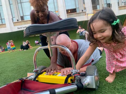 Hettie, a Black woman with curly hair, Petra, a white woman with a shaved head, and tiny community participant, a young white girl, finding sanctuary in color, from Vincent Van Gogh's Asylum Garden painting. All three people reach toward the yellow battery of Petra’s disability scooter. Behind them, the fake grass of an installation extends towards the gray building and tall windows of Lincoln Center. 