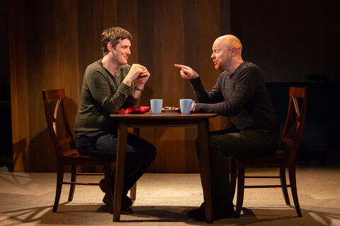 Two performers sit at a table with cups of tea, speaking animatedly.