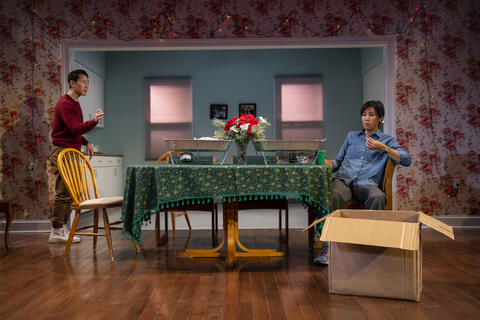 Two performers gather around kitchen table, covered in a floral tablecloth and aluminum serving trays over warmers. There is an open cardboard box downstage.