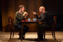 Two performers sit at a table with cups of tea, speaking animatedly.