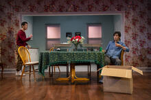 Two performers gather around kitchen table, covered in a floral tablecloth and aluminum serving trays over warmers. There is an open cardboard box downstage.
