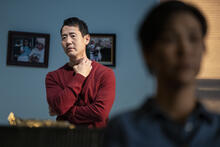 In the foreground, out of focus, a performer sits on a sofa looking out. Another performer watches in the background with a hand placed anxiously at their collar.